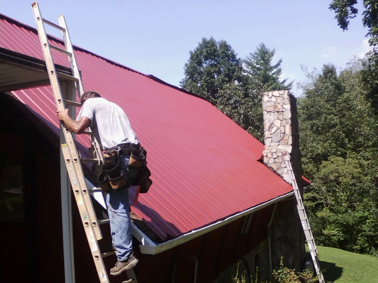 Roofer on a ladder beside a red metal roof and stone chimney — Soffit & Fascia Repair by Roofing Brothers in Neal
